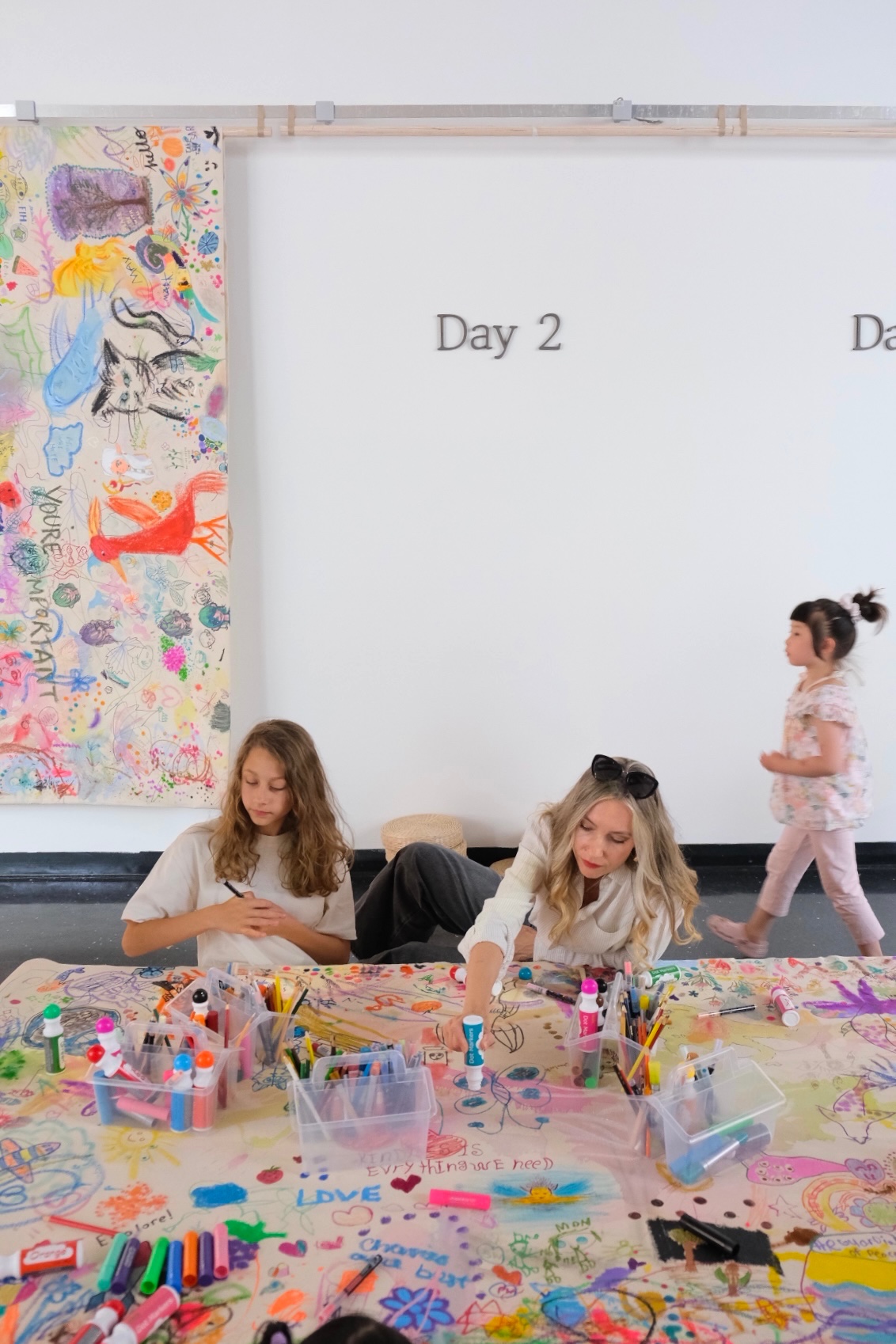 Two visitors make marks together while a child passes the completed Day 2 canvas.